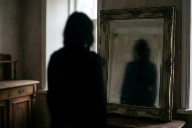 A dark, blurred silhouette stands before an ornate antique mirror in a dim room, reflecting a distorted shadowy figure
